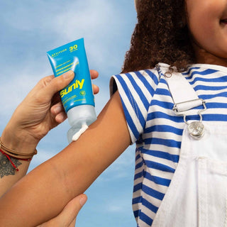 Person applying sunscreen, Attitude Sunly EWG Verified, unscented mineral sunscreen SPF 30 for baby and kids, to another person's arm with a blue sky