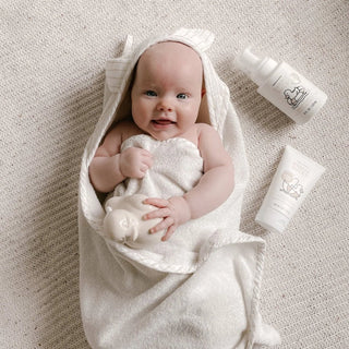 Baby wrapped in a white towel holding a Dabble Ducky with a bottle of Dabble & Dollop baby lotion to the side on a neutral textured background