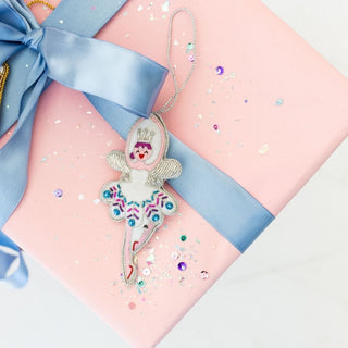 Sugarplum Fairy Ornament resting on a pink gift box tied with blue satin ribbon and scattered with sequins.